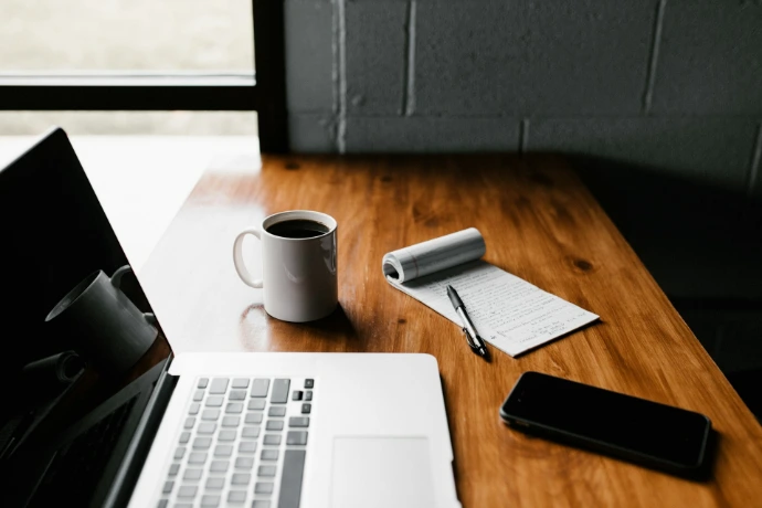 MacBook Pro, cawan seramik putih, dan telefon pintar hitam di atas meja
